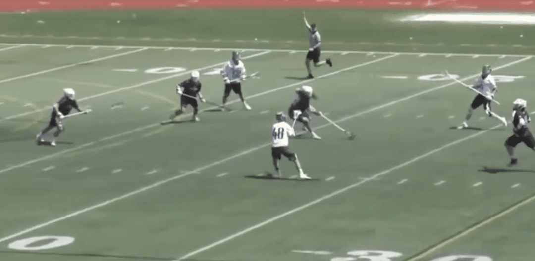 A lacrosse player in a white and light blue jersey defends his goal from the opposing player.